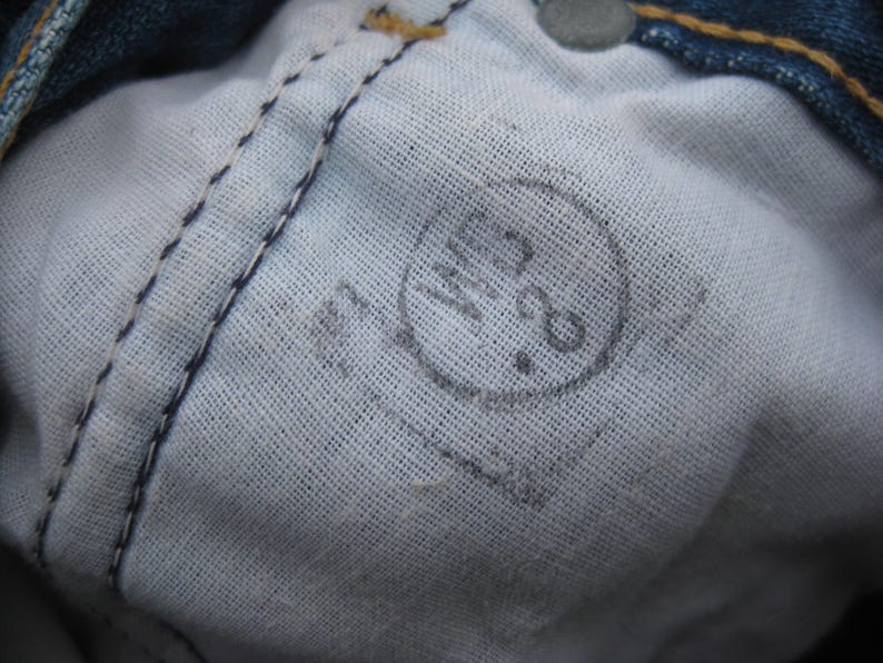May include: Close-up of the inside of denim jeans, showing the white fabric lining and stitching. A faded circular stamp with the numbers "28" is visible. The jeans are blue.