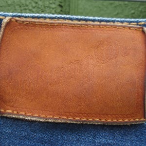 May include: Close-up of a brown leather patch on blue denim jeans. The rectangular patch has a slightly distressed appearance with visible stitching around the edges. The denim fabric is a classic blue color.