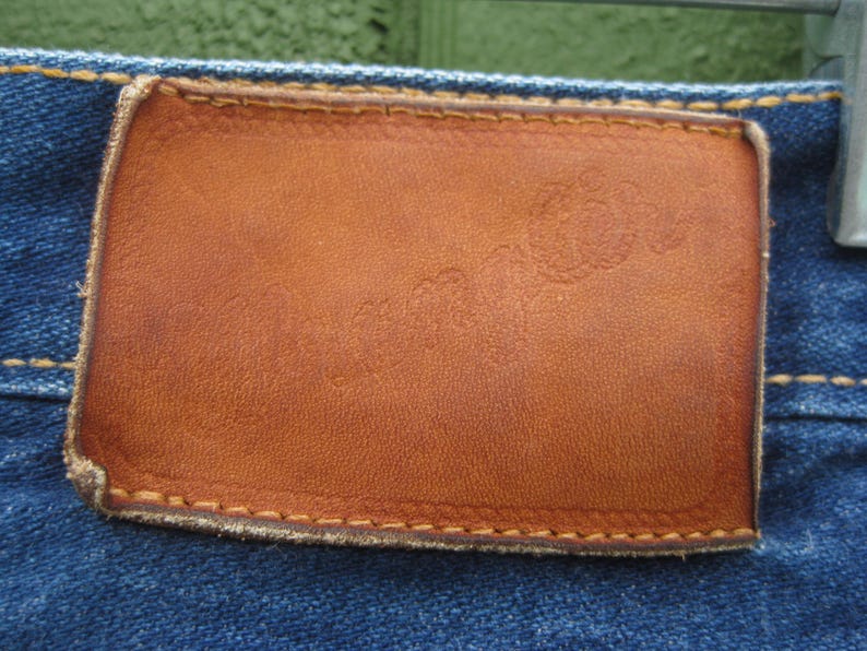 May include: Close-up of a brown leather patch on blue denim jeans. The rectangular patch has a slightly distressed appearance with visible stitching around the edges. The denim fabric is a classic blue color.