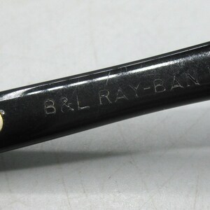 Puede incluir: Primer plano de una patilla de gafas de sol Ray-Ban negras con el texto "B&L RAY-BAN U.S.A." en letras plateadas. La bisagra es visible, con componentes met&aacute;licos plateados. Las gafas de sol son probablemente vintage.