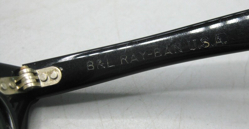 Puede incluir: Primer plano de una patilla de gafas de sol Ray-Ban negras con el texto "B&L RAY-BAN U.S.A." en letras plateadas. La bisagra es visible, con componentes met&aacute;licos plateados. Las gafas de sol son probablemente vintage.