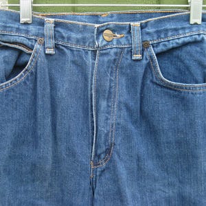 May include: Close-up of blue denim jeans hanging on a metal hanger. The jeans have a button closure, front pockets, and belt loops. The stitching is a contrasting orange color.