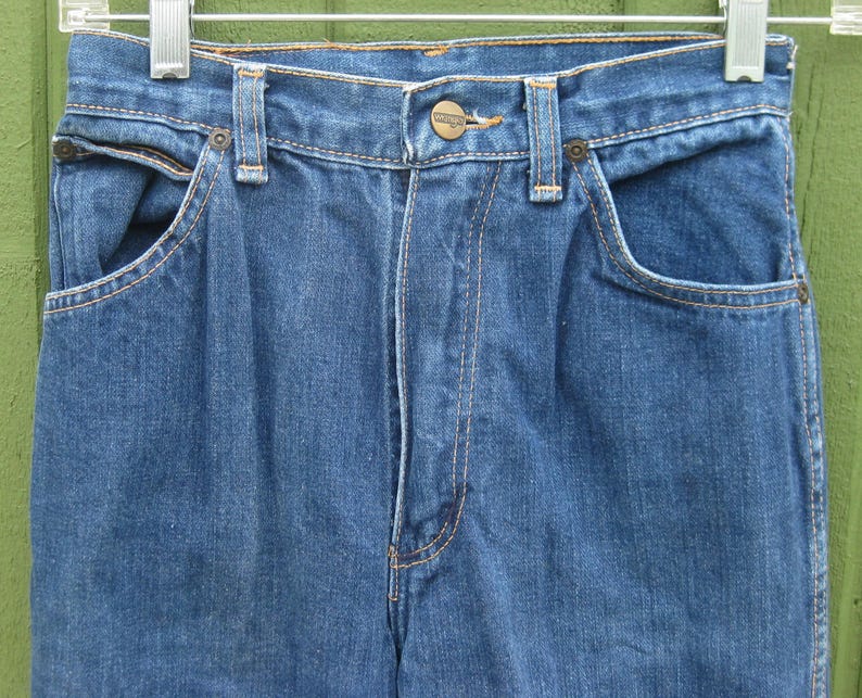 May include: Close-up of blue denim jeans hanging on a metal hanger. The jeans have a button closure, front pockets, and belt loops. The stitching is a contrasting orange color.
