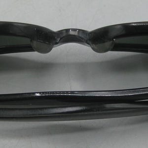 Puede incluir: Gafas de sol negras con un dise&ntilde;o cl&aacute;sico. Las monturas son de color negro brillante y las lentes son oscuras. Las gafas de sol est&aacute;n plegadas, mostrando el interior de las monturas y las lentes. Las patillas son finas y curvas.