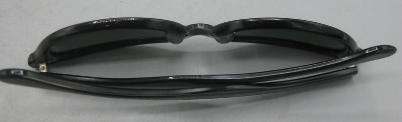 Puede incluir: Gafas de sol negras con un dise&ntilde;o cl&aacute;sico. Las monturas son de color negro brillante y las lentes son oscuras. Las gafas de sol est&aacute;n plegadas, mostrando el interior de las monturas y las lentes. Las patillas son finas y curvas.