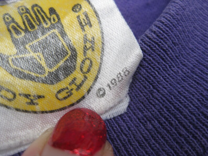 May include: Close-up of a white tag with a yellow and black logo, and the text "Gloves". The tag is attached to a purple knitted fabric. The year "1988" is printed on the tag.