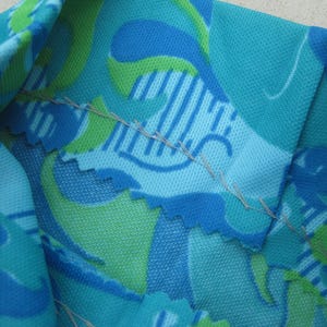 May include: Close-up of a section of fabric with a vibrant, abstract pattern. The design features shades of blue and green, with a textured appearance. The fabric is folded, revealing the stitching along the seams.