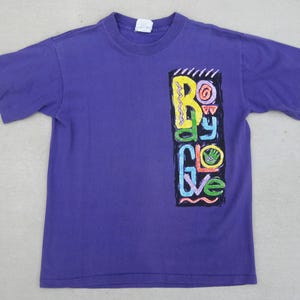 May include: A purple t-shirt with a colorful graphic on the front. The graphic, in a vertical rectangle, features the words "Body Glove" in various bright colors. The shirt has short sleeves and a crew neck.