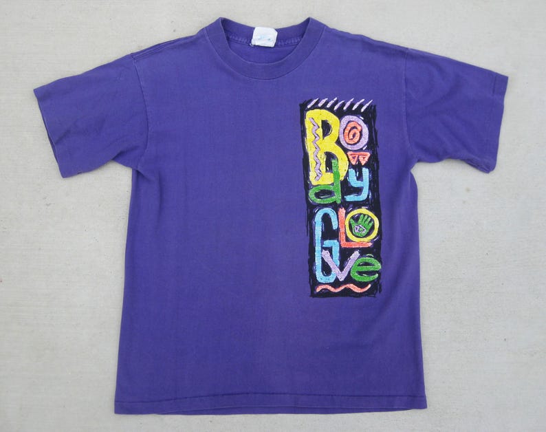 May include: A purple t-shirt with a colorful graphic on the front. The graphic, in a vertical rectangle, features the words "Body Glove" in various bright colors. The shirt has short sleeves and a crew neck.