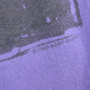 May include: Close-up of a purple fabric with a faded gray graphic design. The text "&copy; 1986 BODY GLOVE" is printed at the bottom. The fabric has a textured appearance.