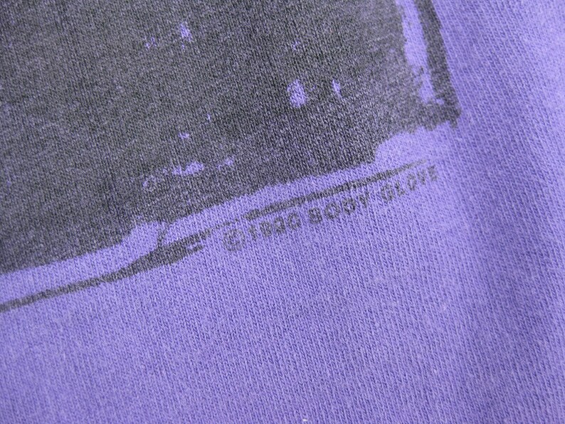May include: Close-up of a purple fabric with a faded gray graphic design. The text "&copy; 1986 BODY GLOVE" is printed at the bottom. The fabric has a textured appearance.