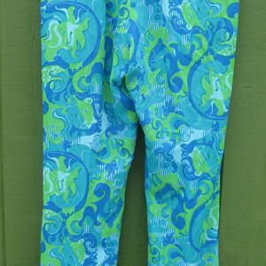 May include: A pair of patterned pants in shades of blue and green, with a repeating abstract design. The pants are hanging on a metal hanger against a green background. The pants have a button and zipper closure.