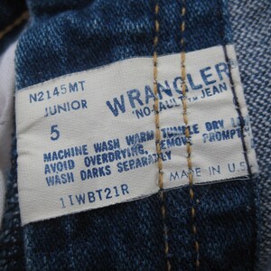 May include: Close-up of a Wrangler jeans tag. The tag is white with blue text, displaying the brand name, size 5 Junior, and washing instructions. The jeans are a classic blue denim.
