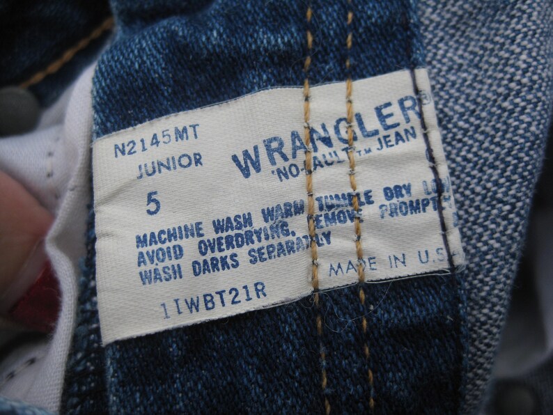 May include: Close-up of a Wrangler jeans tag. The tag is white with blue text, displaying the brand name, size 5 Junior, and washing instructions. The jeans are a classic blue denim.