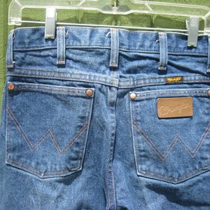 May include: Close-up of the back of blue denim Wrangler jeans hanging on a clear plastic hanger. The jeans have two back pockets with a signature stitching design, copper rivets, and a brown leather patch with the Wrangler logo.