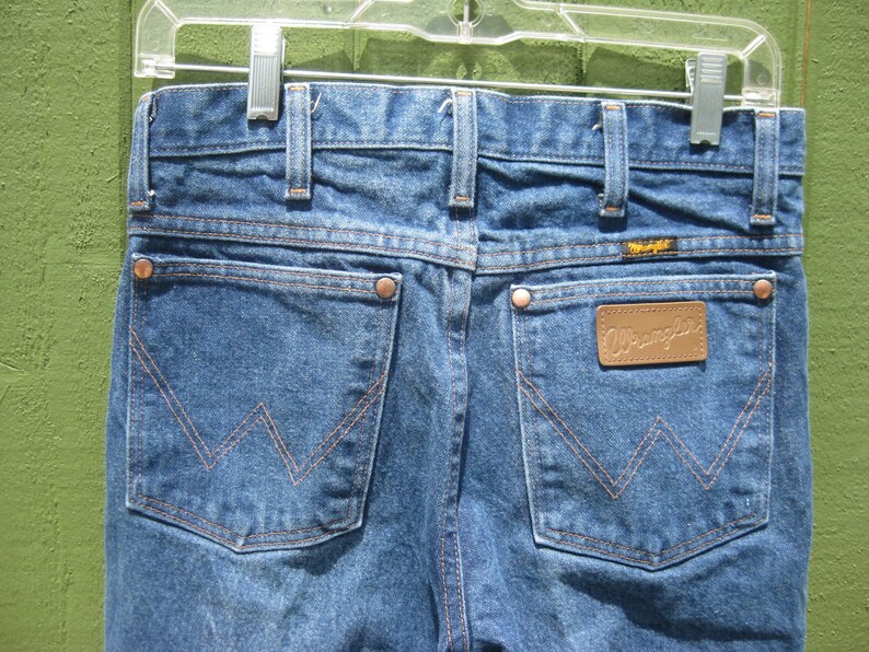 May include: Close-up of the back of blue denim Wrangler jeans hanging on a clear plastic hanger. The jeans have two back pockets with a signature stitching design, copper rivets, and a brown leather patch with the Wrangler logo.