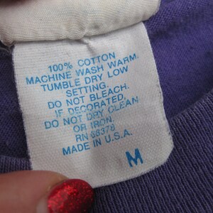 May include: Close-up of a white tag with blue text on a purple shirt. The tag reads "100% COTTON, MACHINE WASH WARM, TUMBLE DRY LOW, DO NOT BLEACH, MADE IN U.S.A." The tag also has the size "M".