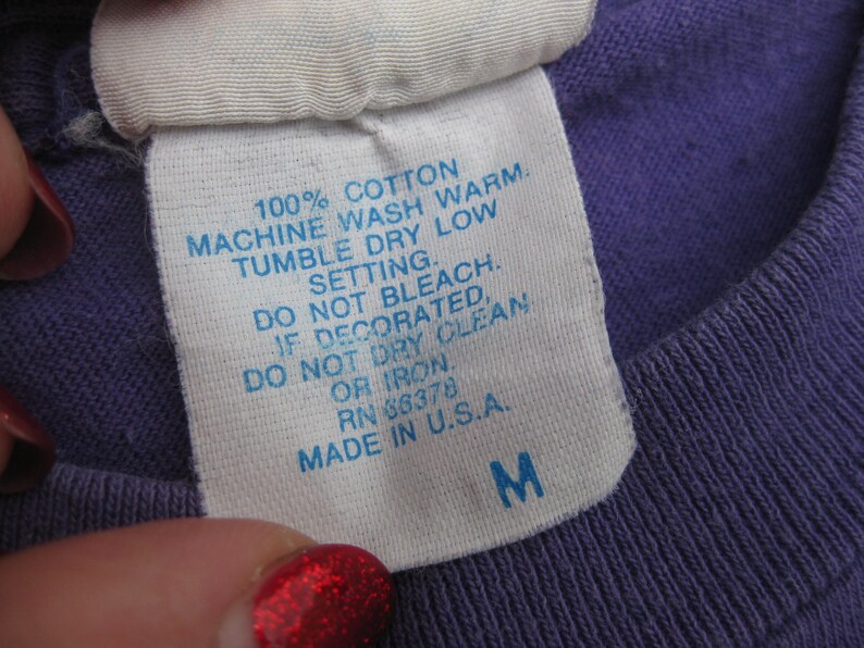May include: Close-up of a white tag with blue text on a purple shirt. The tag reads "100% COTTON, MACHINE WASH WARM, TUMBLE DRY LOW, DO NOT BLEACH, MADE IN U.S.A." The tag also has the size "M".