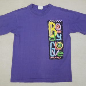 May include: A purple t-shirt with a crew neck and short sleeves. The shirt features a colorful graphic on the front with the words "Body Glove" in a vertical design. The graphic includes yellow, green, blue, and pink elements.