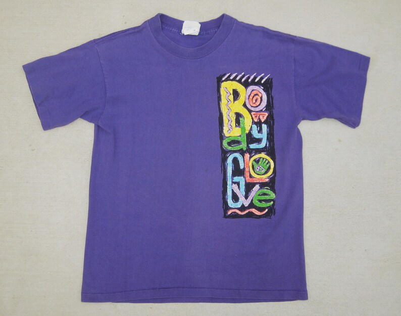 May include: A purple t-shirt with a crew neck and short sleeves. The shirt features a colorful graphic on the front with the words "Body Glove" in a vertical design. The graphic includes yellow, green, blue, and pink elements.