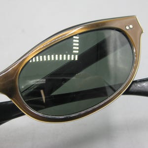 Puede incluir: Primer plano de gafas de sol vintage con montura marr&oacute;n y lentes verde oscuro. Las gafas de sol tienen una forma cl&aacute;sica de ojo de gato con dos peque&ntilde;as remaches en las esquinas superiores de la montura. Las patillas son negras.