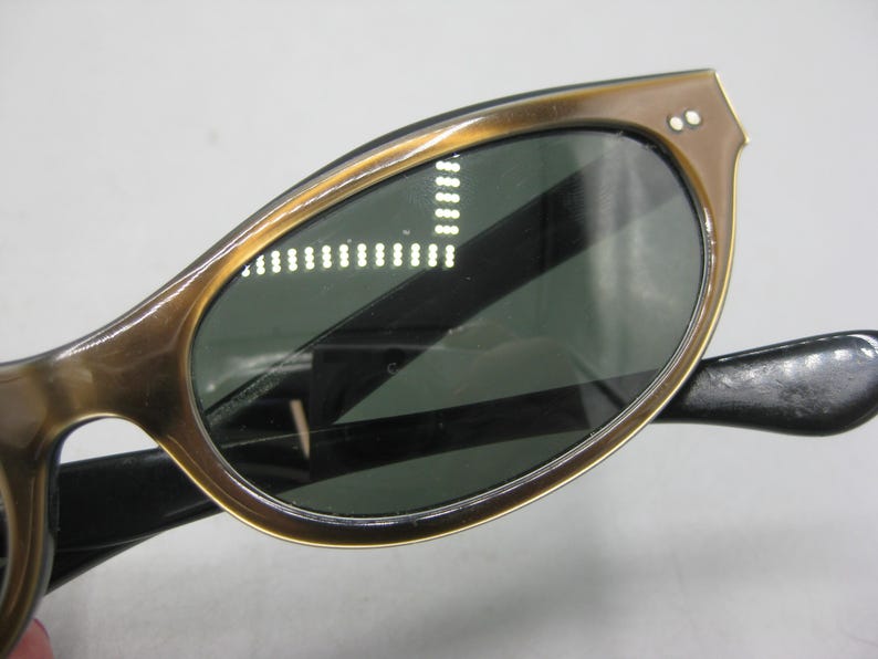Puede incluir: Primer plano de gafas de sol vintage con montura marr&oacute;n y lentes verde oscuro. Las gafas de sol tienen una forma cl&aacute;sica de ojo de gato con dos peque&ntilde;as remaches en las esquinas superiores de la montura. Las patillas son negras.