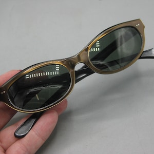 Puede incluir: Gafas de sol vintage de ojo de gato con montura dorada y lentes oscuras. Las patillas son negras. Las gafas de sol se sostienen en una mano.