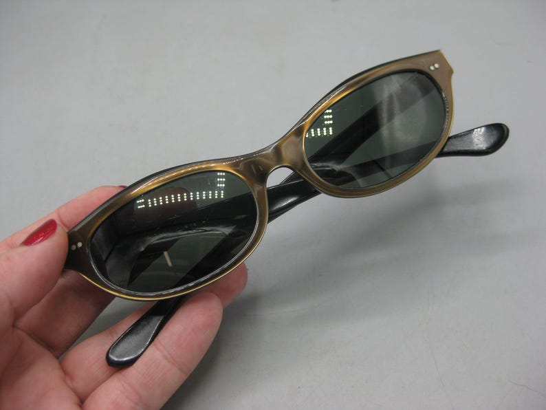 Puede incluir: Gafas de sol vintage de ojo de gato con montura dorada y lentes oscuras. Las patillas son negras. Las gafas de sol se sostienen en una mano.