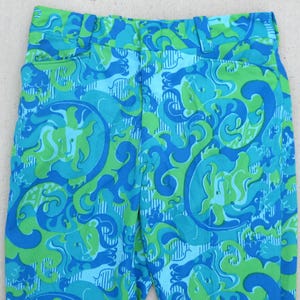 May include: Close-up of vintage pants with a vibrant, swirling pattern in shades of blue and green. The fabric features a repeating design of abstract shapes and forms, creating a psychedelic aesthetic. The pants have a classic cut with a waistband and pockets.