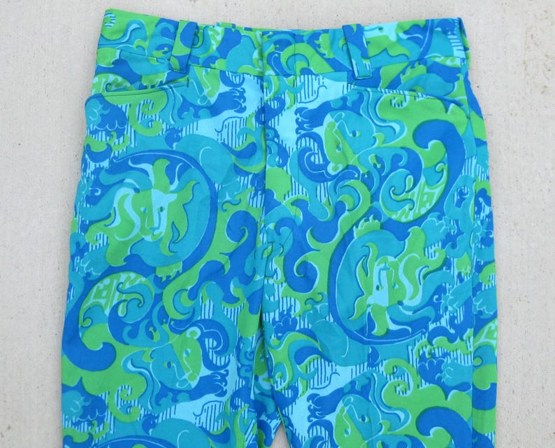 May include: Close-up of vintage pants with a vibrant, swirling pattern in shades of blue and green. The fabric features a repeating design of abstract shapes and forms, creating a psychedelic aesthetic. The pants have a classic cut with a waistband and pockets.
