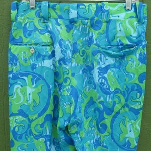 May include: A pair of vintage trousers with a vibrant, swirling pattern in shades of turquoise, blue, and lime green. The trousers feature a classic design with belt loops, pockets, and a button closure. The fabric has a slight sheen.