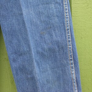 May include: Close-up of blue denim jeans. The jeans have a classic design with visible stitching along the seams and a slight discoloration on the fabric. The background is a solid green color.
