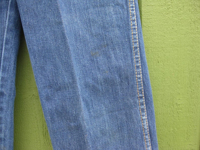 May include: Close-up of blue denim jeans. The jeans have a classic design with visible stitching along the seams and a slight discoloration on the fabric. The background is a solid green color.