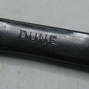 Puede incluir: Primer plano de un objeto gris oscuro con la palabra "DUNE" grabada en su superficie. El objeto parece ser de metal y tiene un acabado ligeramente texturizado. La iluminaci&oacute;n resalta el texto grabado.