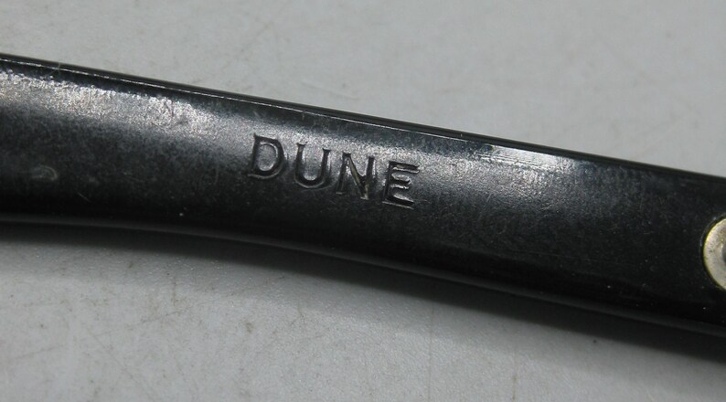 Puede incluir: Primer plano de un objeto gris oscuro con la palabra "DUNE" grabada en su superficie. El objeto parece ser de metal y tiene un acabado ligeramente texturizado. La iluminaci&oacute;n resalta el texto grabado.