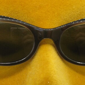 Puede incluir: Gafas de sol marrones oscuras con forma de ojo de gato y un brillo sutil en la parte superior de la montura. Las lentes son de color verde oscuro y las gafas tienen un dise&ntilde;o cl&aacute;sico y retro. Las gafas se muestran en una cabeza de maniqu&iacute; amarilla.