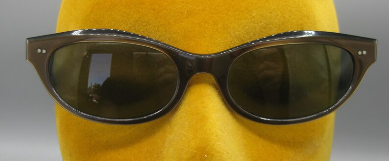 Puede incluir: Gafas de sol marrones oscuras con forma de ojo de gato y un brillo sutil en la parte superior de la montura. Las lentes son de color verde oscuro y las gafas tienen un dise&ntilde;o cl&aacute;sico y retro. Las gafas se muestran en una cabeza de maniqu&iacute; amarilla.