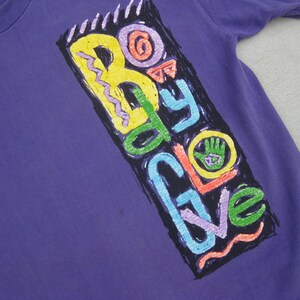 May include: A purple t-shirt with a colorful graphic that reads "Body Glove." The graphic features a hand with a green palm and the brand name in various colors, including yellow, green, blue, and orange. The shirt has short sleeves.