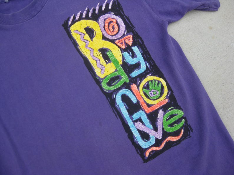 May include: A purple t-shirt with a colorful graphic that reads "Body Glove." The graphic features a hand with a green palm and the brand name in various colors, including yellow, green, blue, and orange. The shirt has short sleeves.
