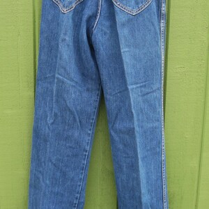 May include: Blue denim jeans with a classic design. Features include two back pockets with decorative stitching, a brown leather patch, and white stitching along the seams. The jeans are hanging on a white hanger.