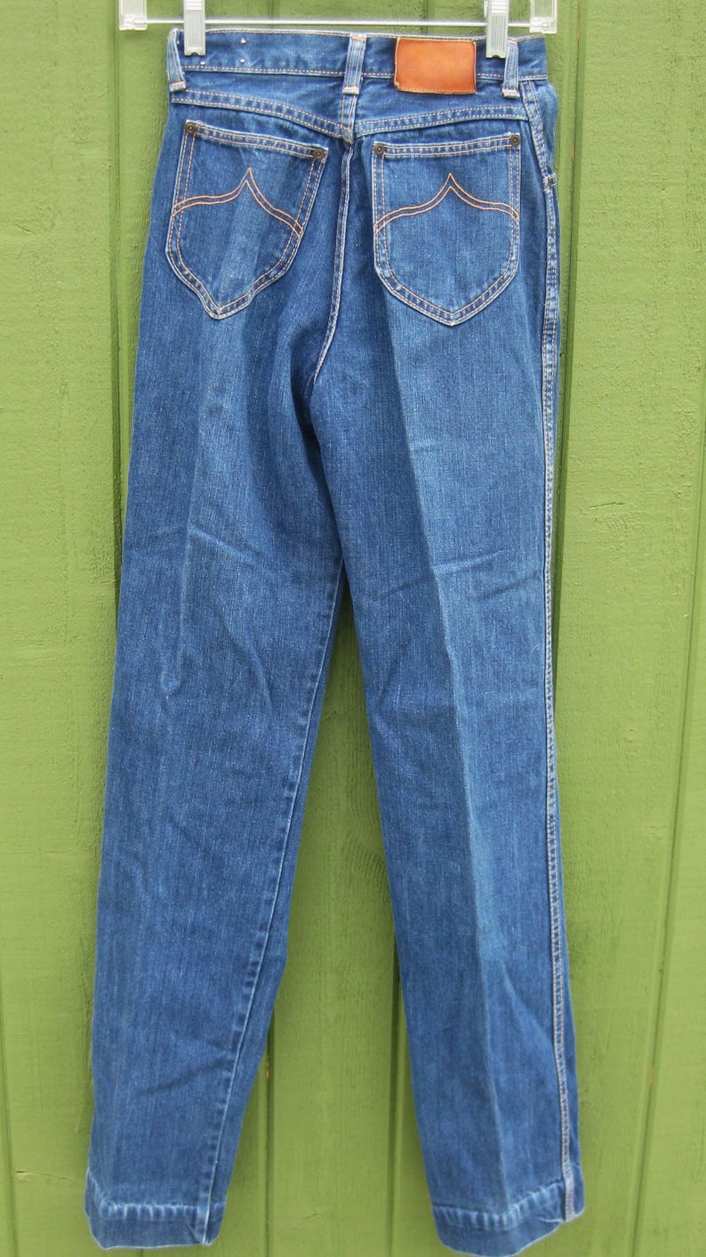 May include: Blue denim jeans with a classic design. Features include two back pockets with decorative stitching, a brown leather patch, and white stitching along the seams. The jeans are hanging on a white hanger.