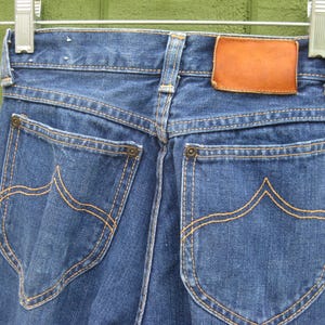 May include: Close-up of the back of blue denim jeans hanging on a hanger. The jeans have two back pockets with decorative stitching in a contrasting orange thread. A brown leather patch is visible at the waistband.