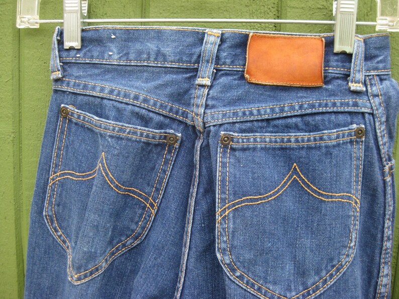 May include: Close-up of the back of blue denim jeans hanging on a hanger. The jeans have two back pockets with decorative stitching in a contrasting orange thread. A brown leather patch is visible at the waistband.