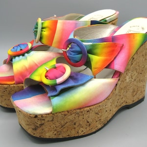 May include: A pair of rainbow-colored wedge sandals with cork soles. The sandals feature a vibrant spectrum of colors and large decorative buckles. The platform heels are approximately 4 inches high.