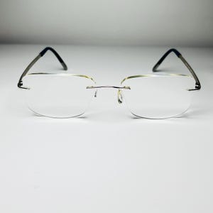 May include: Rimless eyeglasses with clear lenses and thin, silver-toned metal frames. The temples are dark gray. The glasses are designed for a minimalist aesthetic.