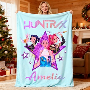May include: A light blue blanket featuring the text "HUNTRAX" and the name "Amelia". The blanket has a graphic of three cartoon women inside a purple star, with a blue creature. The blanket is held up by a person.
