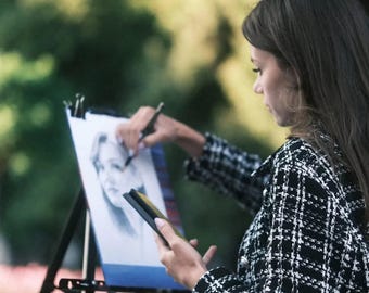 May include: A woman in a black and white checkered jacket is drawing a portrait on a canvas easel. She is using a pencil to draw the portrait.