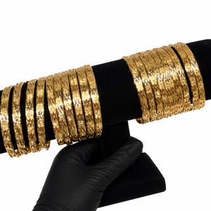 May include: A set of gold-colored bangles featuring a repeating star-like etched pattern. The bangles are arranged in two groups on a black cylindrical display stand, showcasing a reflective metallic finish and consistent circular design throughout.