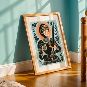 May include: A framed art print featuring a stylized illustration of a knight in black armor holding a sword and cross. The knight has a halo and is set against a light blue background with floral accents. The artwork is in a wooden frame.
