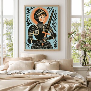 May include: A framed art print of a medieval knight in black armor, holding a sword. The artwork features a light blue background with an orange halo and decorative black foliage. The print is displayed above a bed with beige bedding.